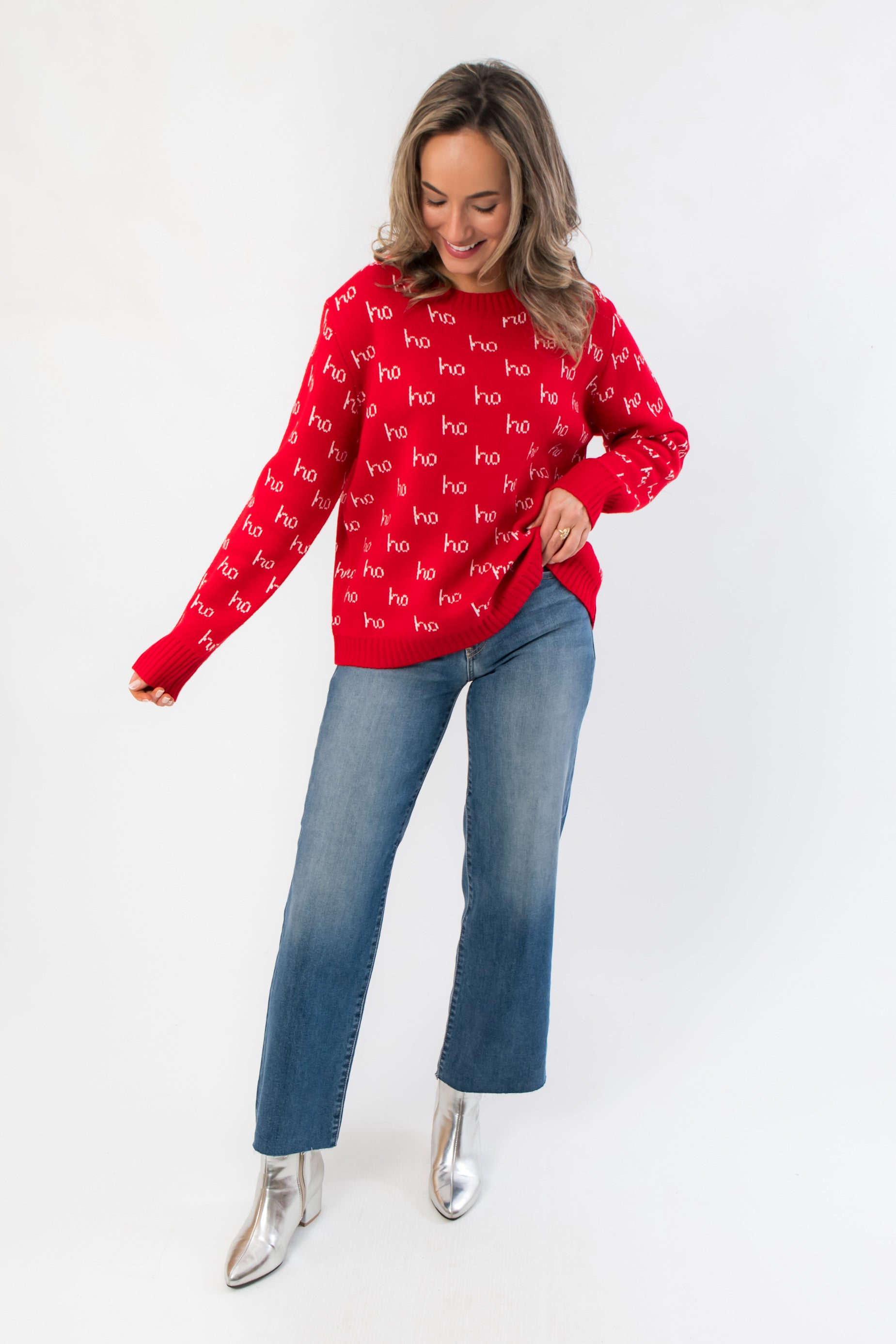 Boutique holiday style red Christmas sweater featuring festive 'ho' print, paired with flared jeans and silver ankle boots.