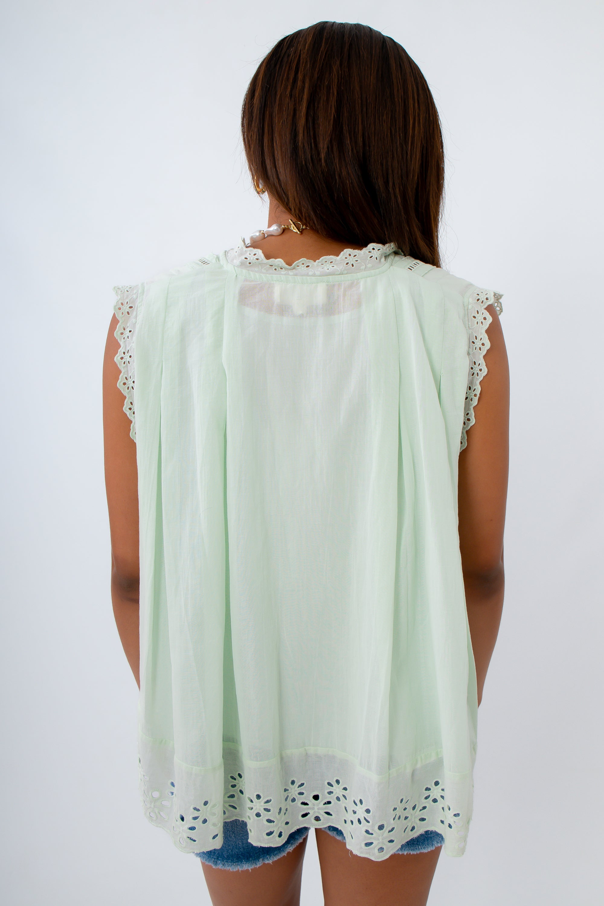 A woman viewed from behind wearing a sleeveless mint green blouse with white eyelet lace trim at the neckline and hem, paired with denim shorts against a plain white background.