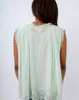 A woman viewed from behind wearing a sleeveless mint green blouse with white eyelet lace trim at the neckline and hem, paired with denim shorts against a plain white background.