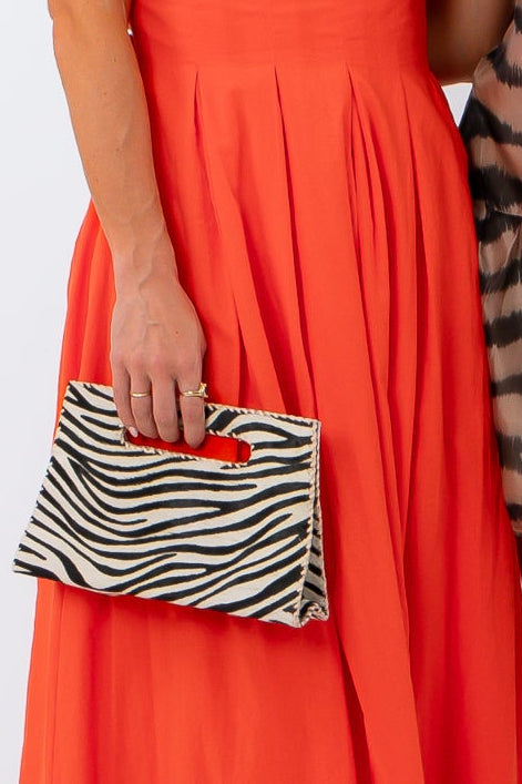 Two women posing together against a white studio background. The woman on the left wears a bright orange sleeveless maxi dress and holds a zebra-print clutch.