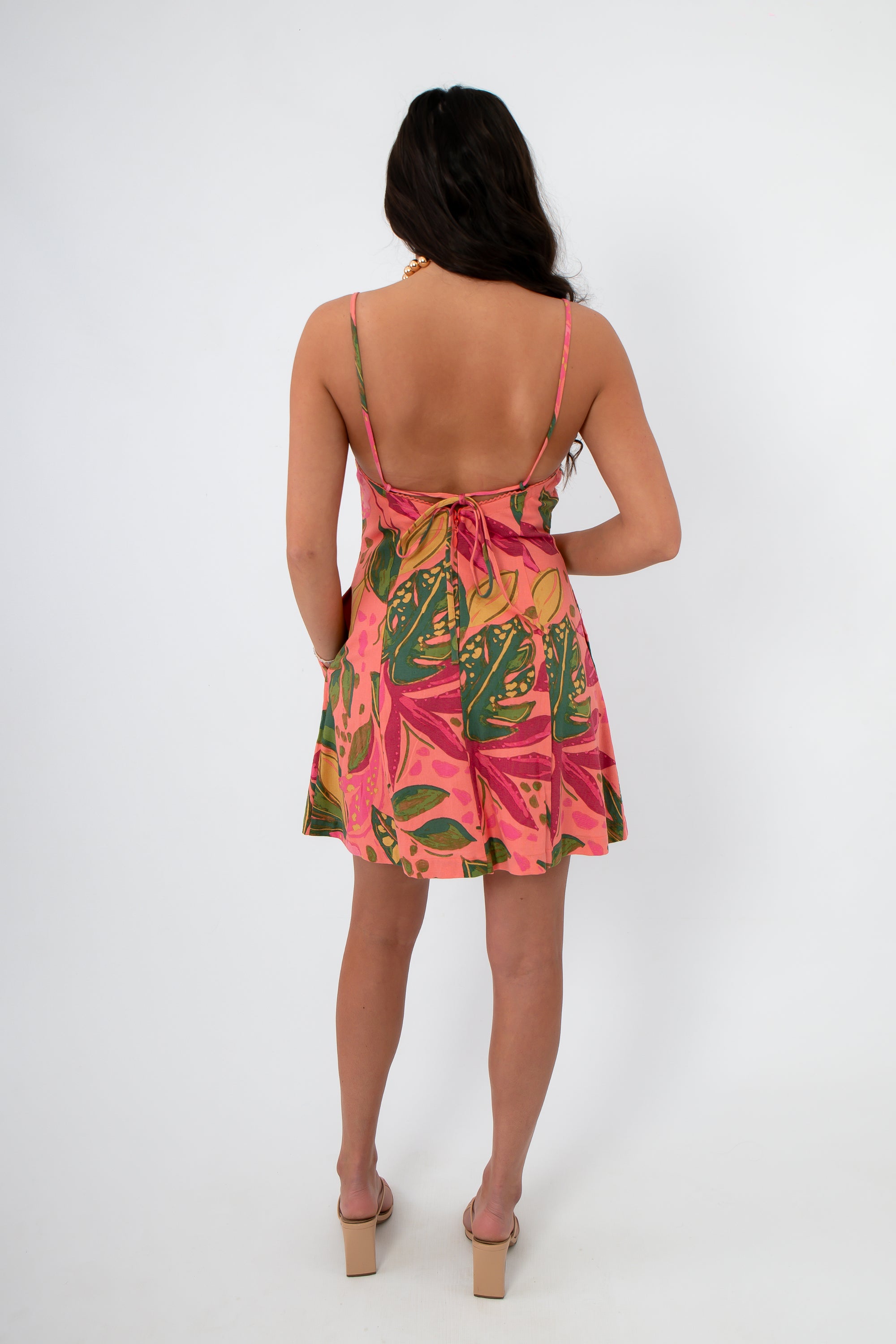 Back view of Cabana Nights Tropical Printed Mini Dress in coral with pink and green leaf pattern, featuring open back and tie detail.