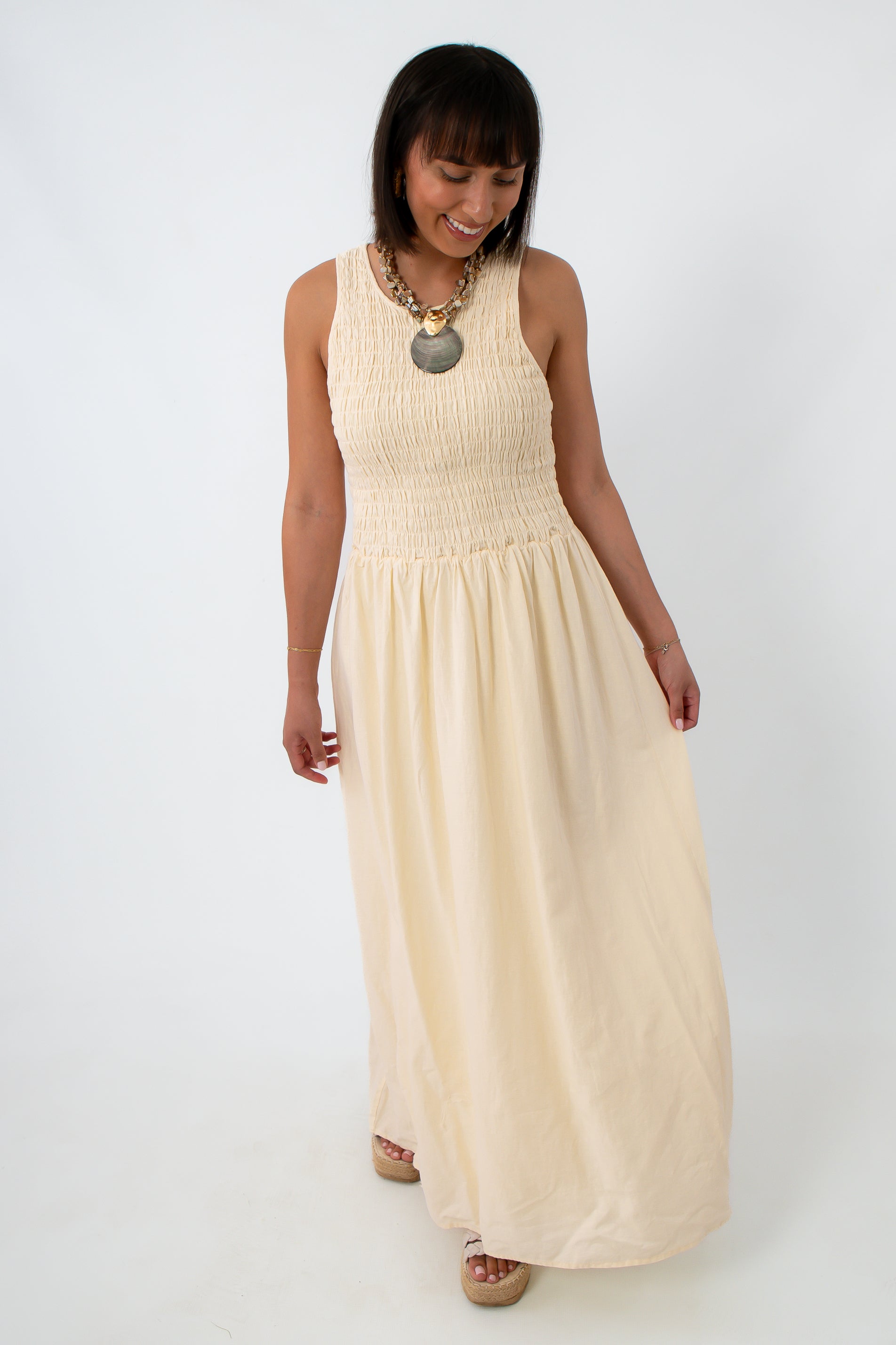 Side view of woman in cream maxi dress with smocked top and flowing skirt, smiling and looking down