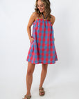 Back view of woman in a red and blue plaid mini dress with adjustable straps, standing with hands in pockets, white background