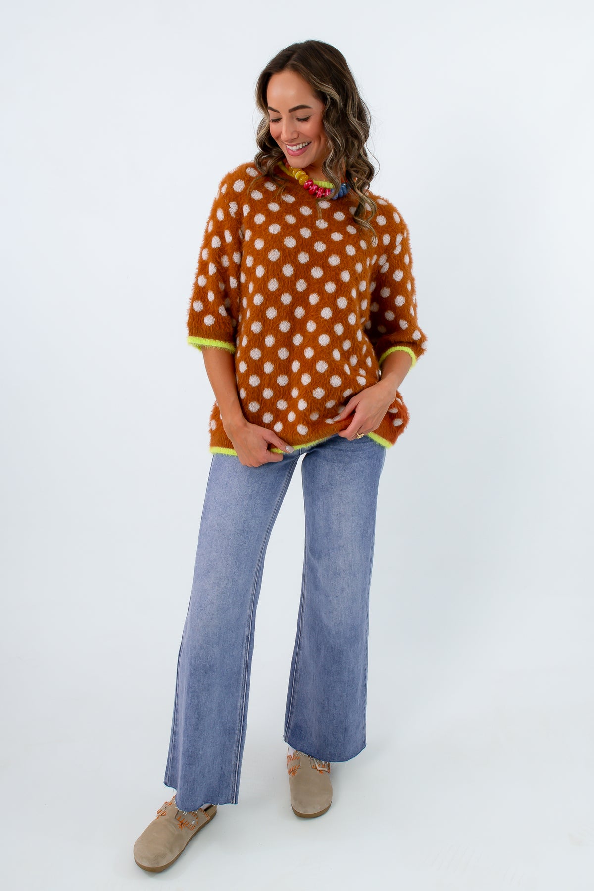 A woman modeling a rust-orange knit sweater with white polka dots and bright yellow-green trim, paired with wide-leg blue jeans and tan shoes, against a plain white studio background.