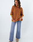 A woman modeling a rust-orange knit sweater with white polka dots and bright yellow-green trim, paired with wide-leg blue jeans and tan shoes, against a plain white studio background.