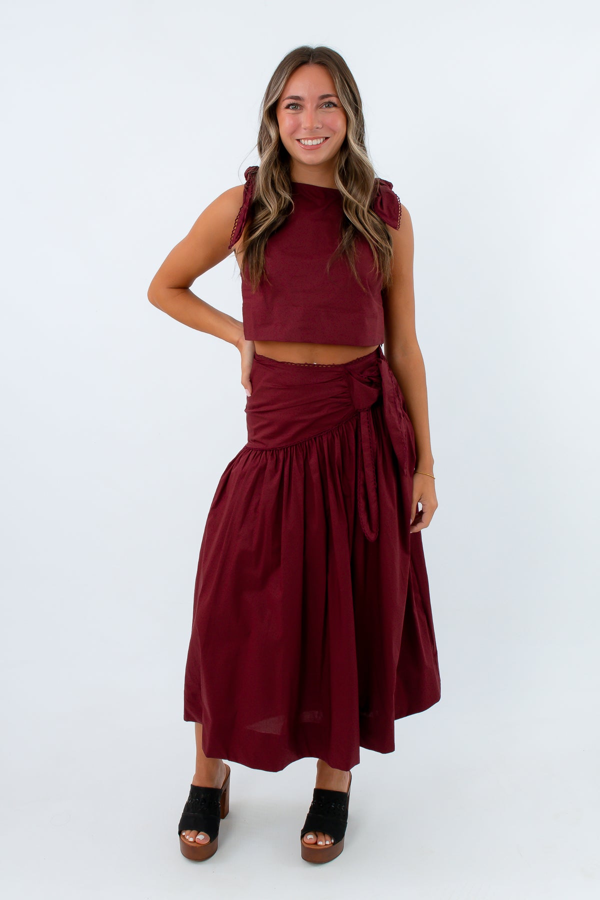 A woman modeling a matching burgundy two-piece set consisting of a sleeveless crop top with shoulder ties and a high-waisted wrap midi skirt, paired with black platform sandals, against a plain white studio background.