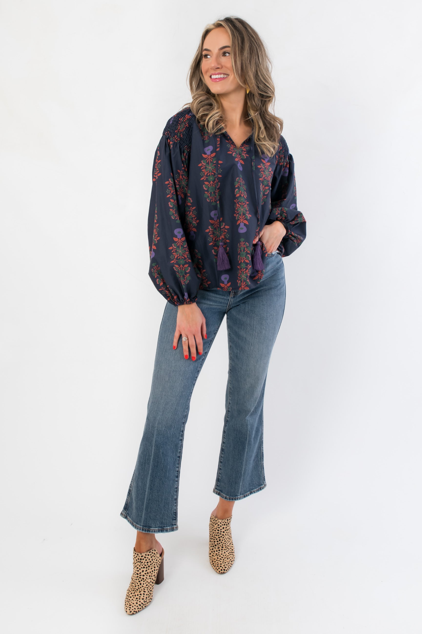 Full-body view of At Long Last Navy Long Sleeve Blouse featuring floral balloon sleeves and tassel ties, paired with jeans and heeled mules.
