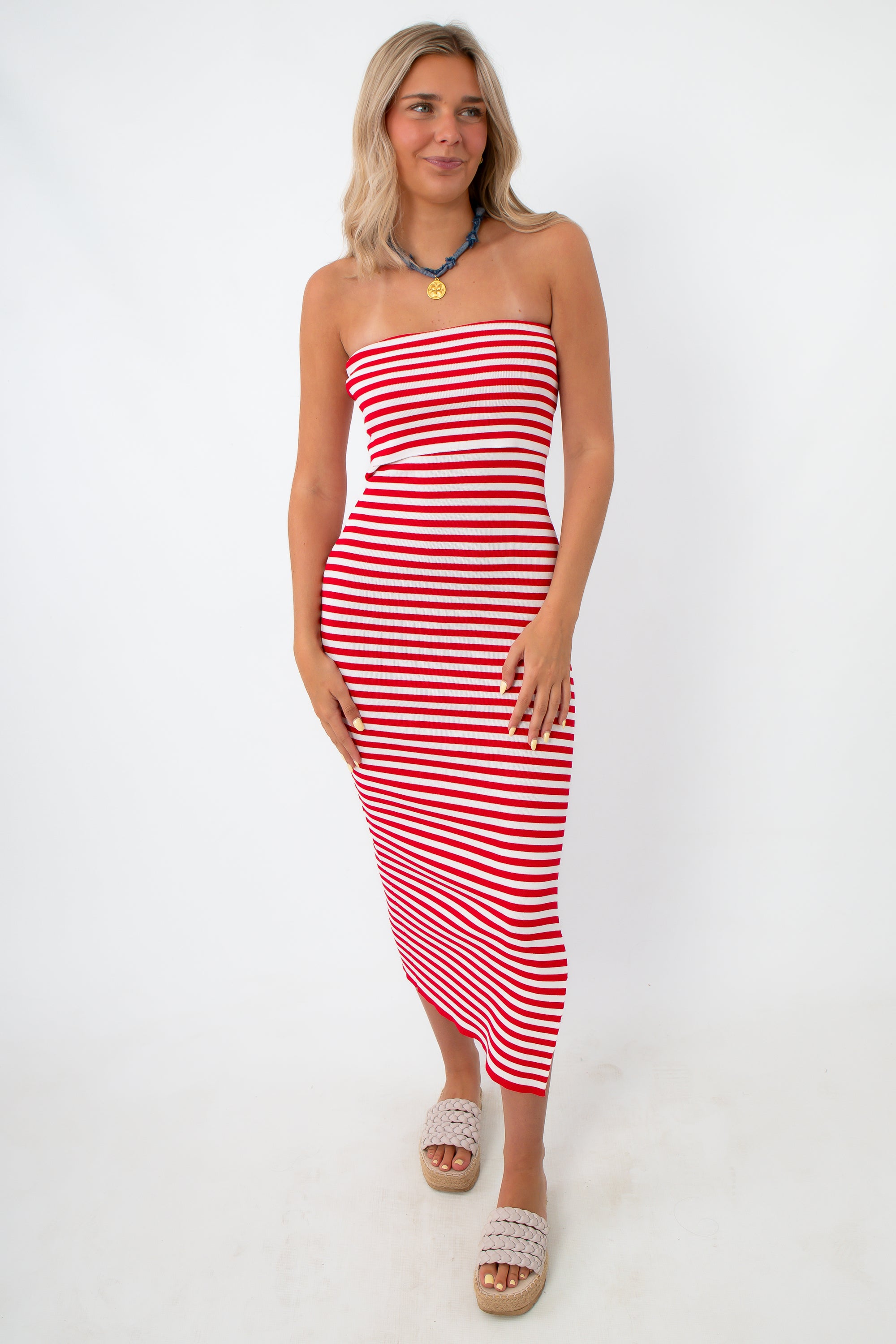 Side view of woman in red and white striped strapless knit midi dress with side slit, wearing beige platform sandals and a blue necklace, standing against white background