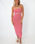 Side view of woman in red and white striped strapless knit midi dress with side slit, wearing beige platform sandals and a blue necklace, standing against white background