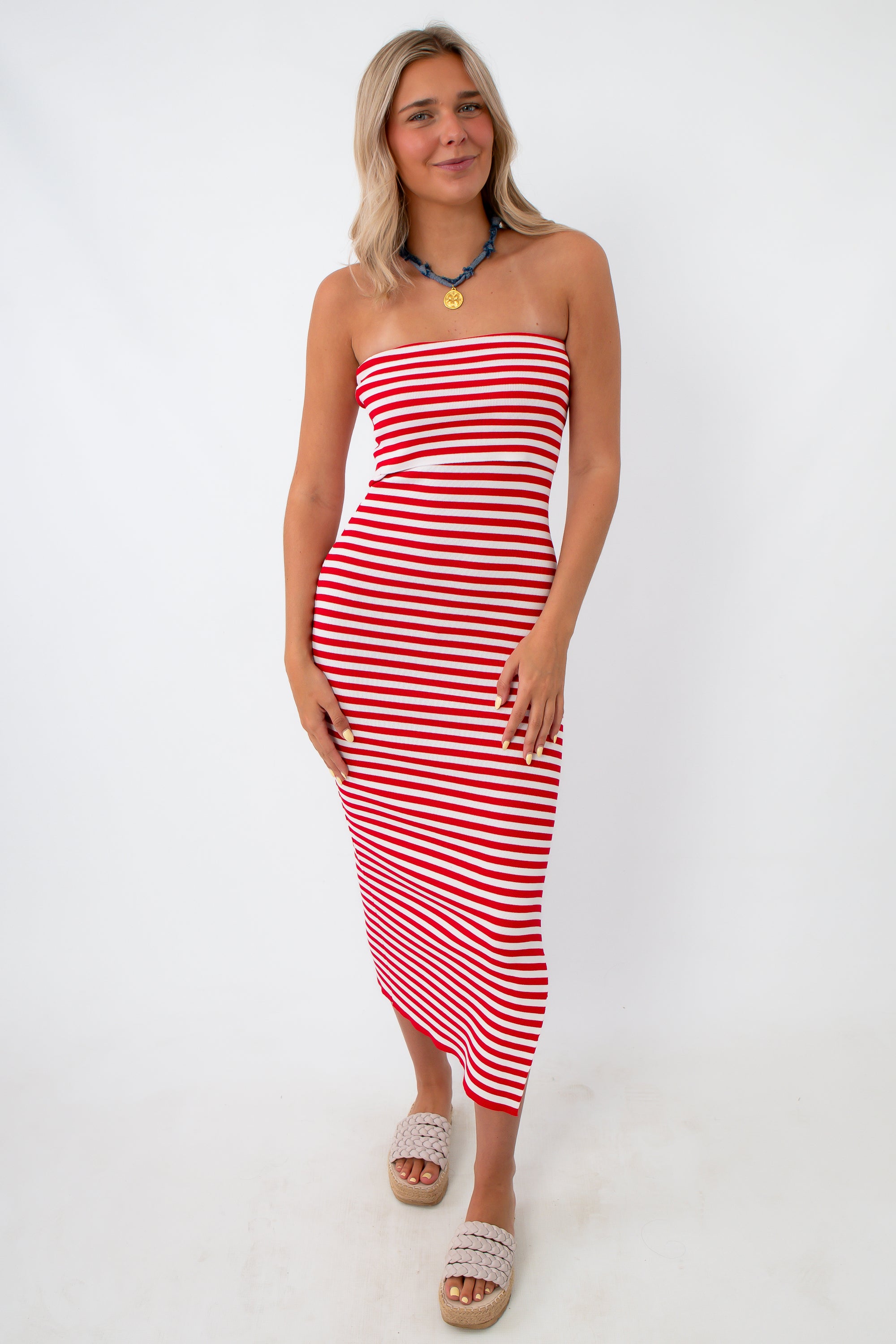 Woman in red and white striped strapless knit midi dress with side slit, wearing beige platform sandals and a blue necklace, standing against white background