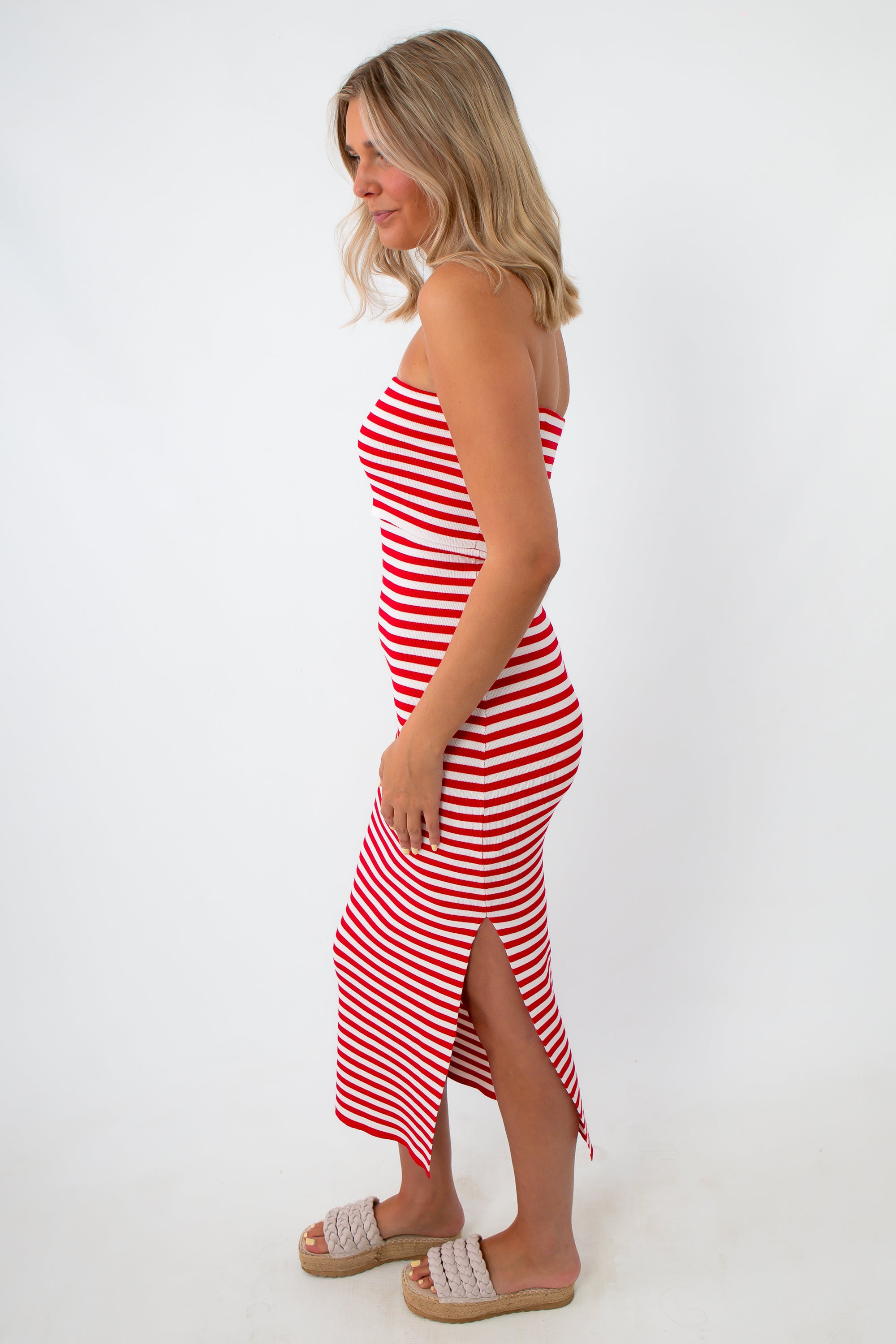 Back view of woman in red and white striped strapless knit midi dress with side slit, wearing beige platform sandals, standing against white background