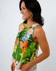 Back view of woman with dark wavy hair, wearing tropical print sleeveless blouse, holding straw bag, white background