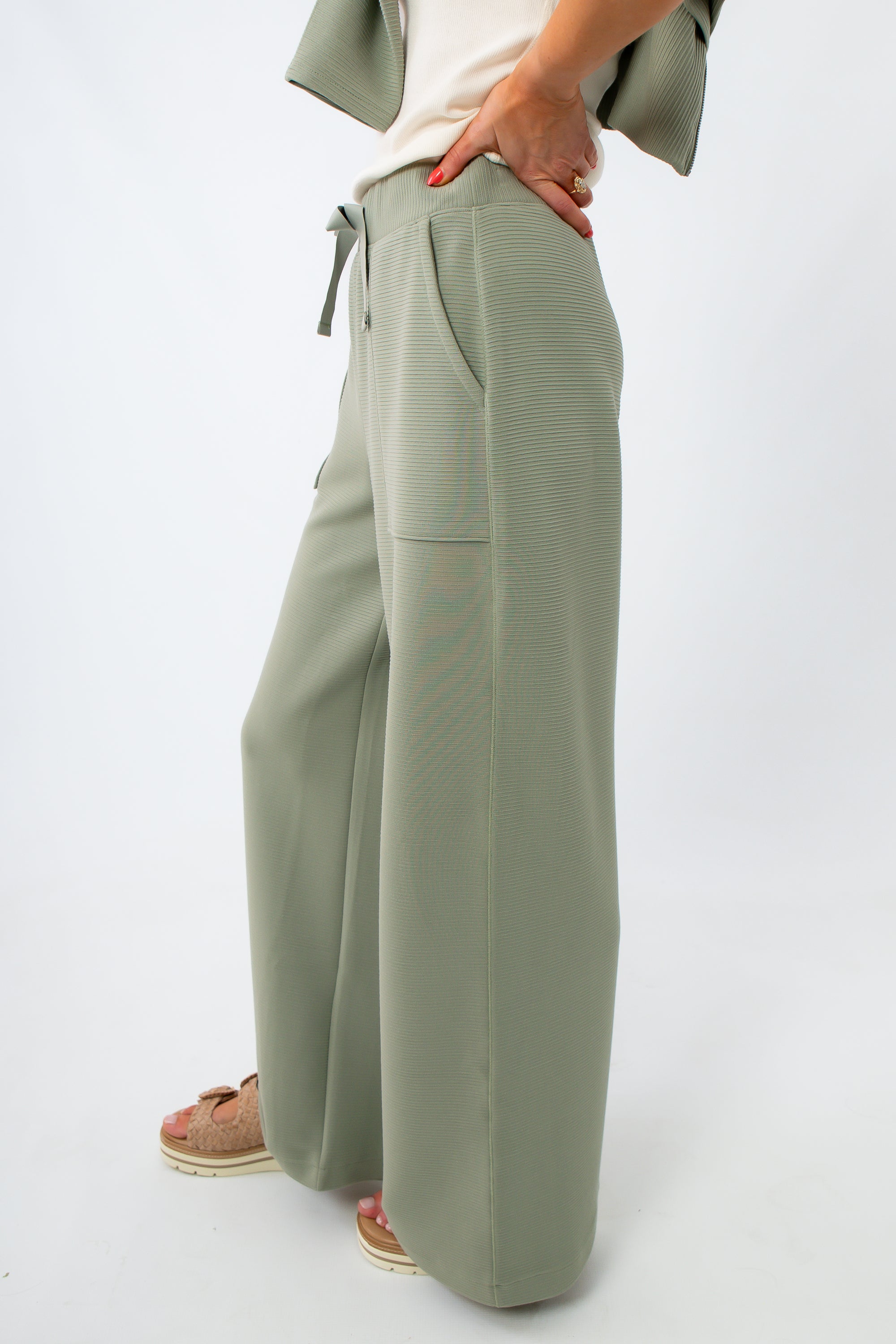 Woman in sage green lounge pants and white top, smiling and posing sideways