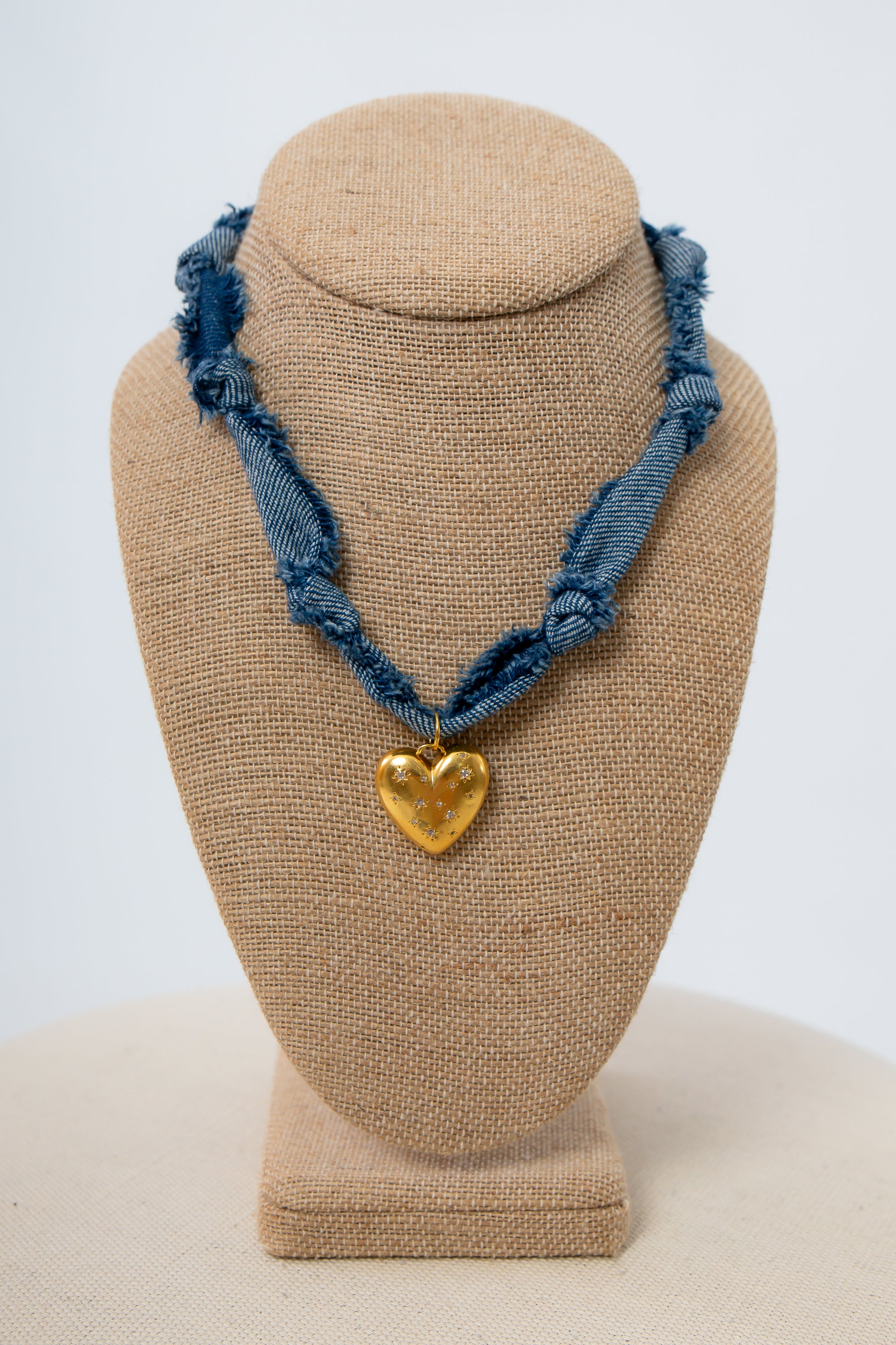 Heart of Gold Denim Bandana Necklace Blue braided necklace with a gold heart pendant on a beige bust against a white background