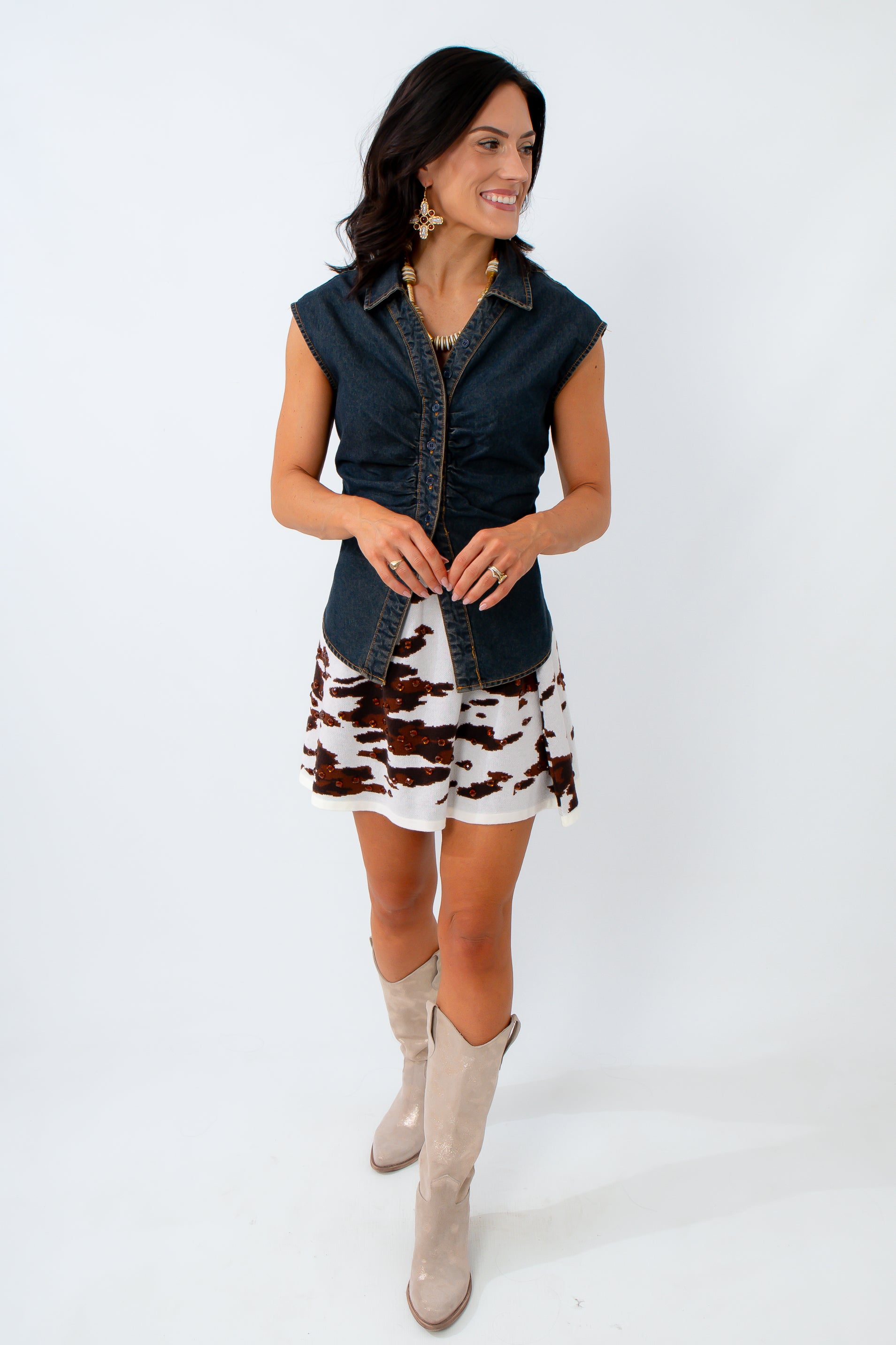 Woman wearing a denim vest over a patterned dress with beige boots on a white background
