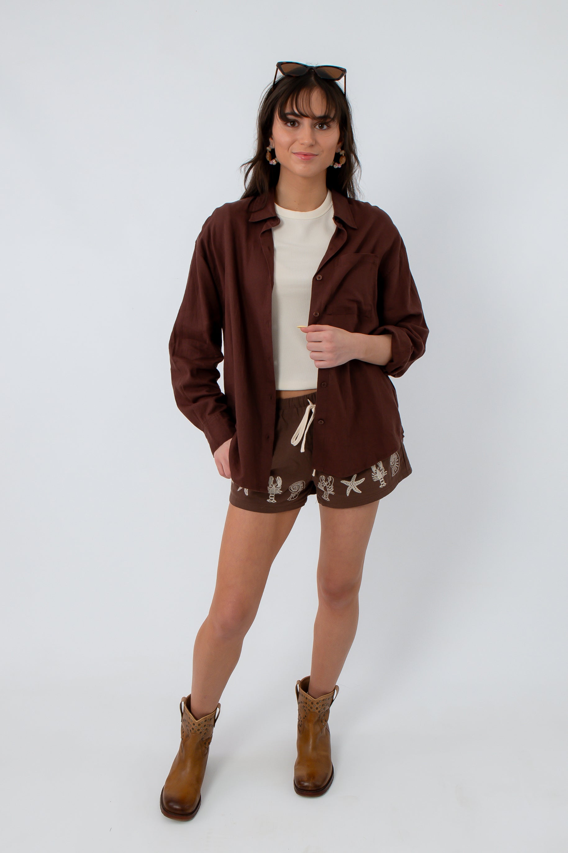 Women’s brown button-up shirt with long sleeves, layered over a white top, paired with embroidered shorts and cowboy boots