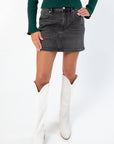 Daze Spade Malibu Mid Waist Mini Skirt in washed black denim with A-line silhouette and front button detail, styled with white boots.