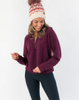 Spanx Plum Berry Air Essentials Half Zip Sweatshirt for women in a rich plum color, styled with black leggings and a knit beanie.