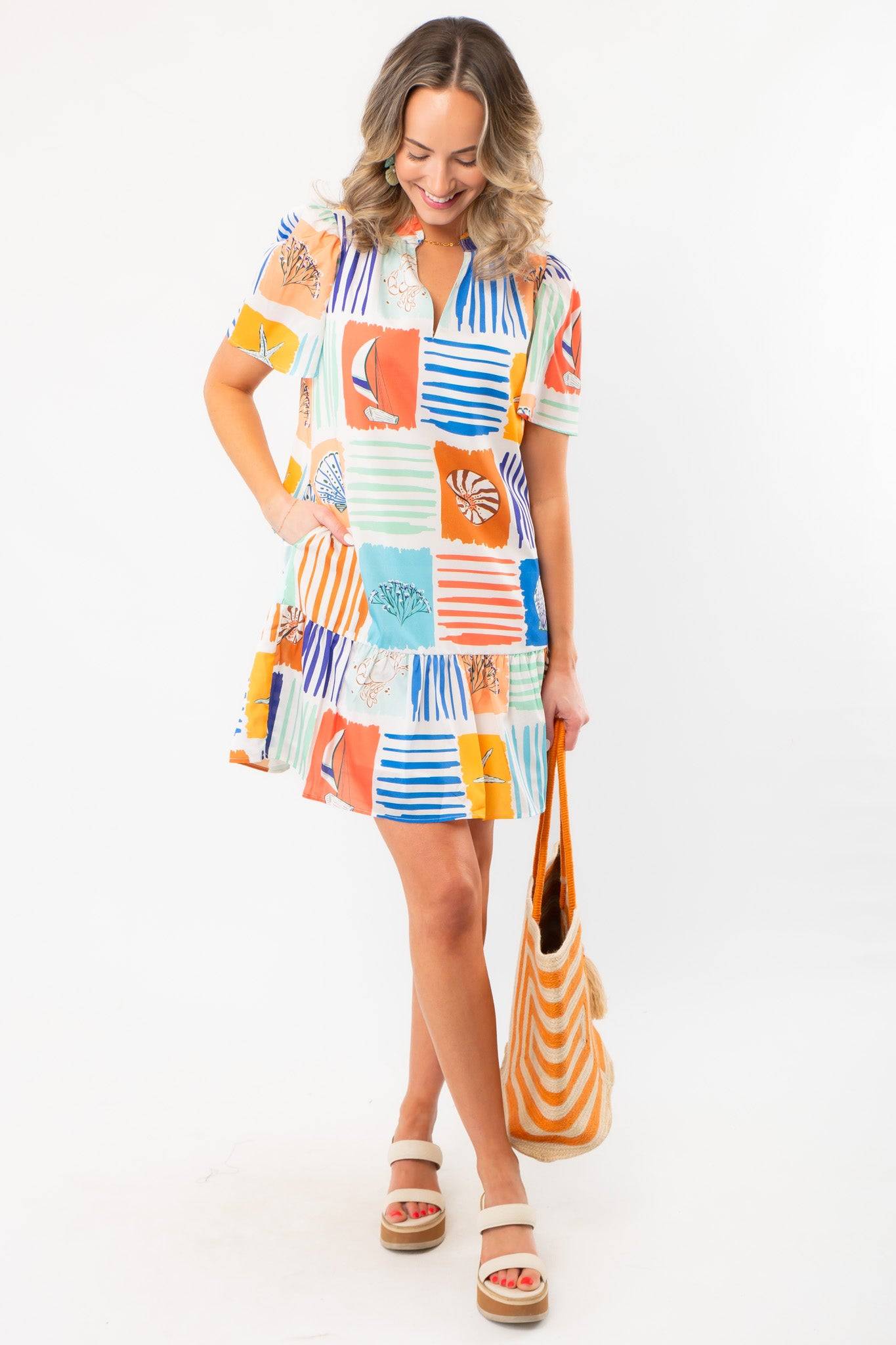 Model wearing THML At Sea Nautical Printed Dress with beach-themed prints and pockets, styled with a straw tote for a casual summer look.