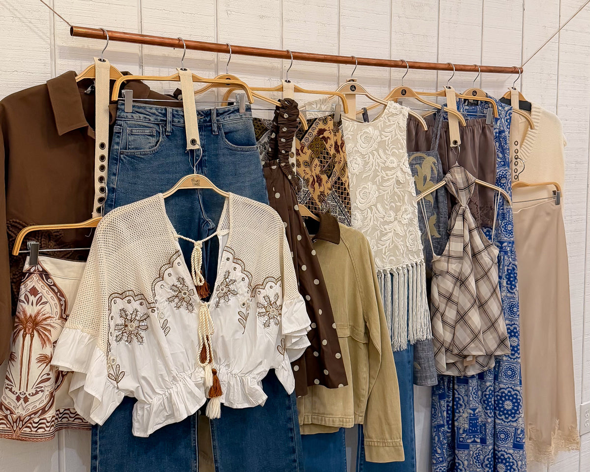 wild mabel pawleys island boutique A clothing rack display in a boutique featuring a variety of women's apparel, including blue jeans, a white embroidered blouse, a brown jacket, and patterned dresses, set against a white wood-paneled wall.