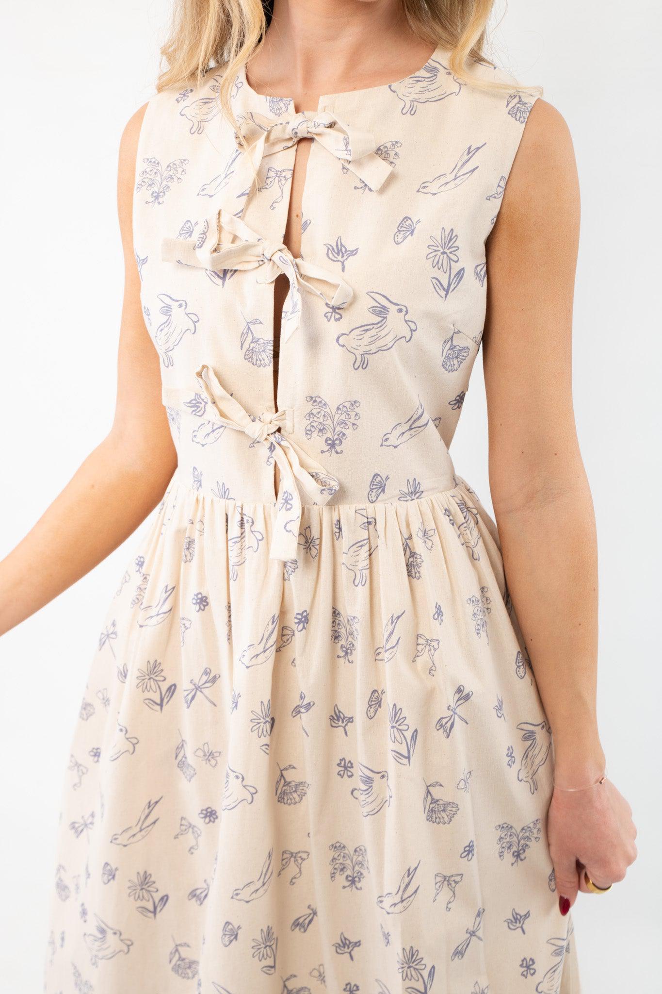 Close-up of the Hop To It Beige Bunny Pattern Midi Dress featuring a playful blue bunny and floral print with a triple bow closure on the bust.