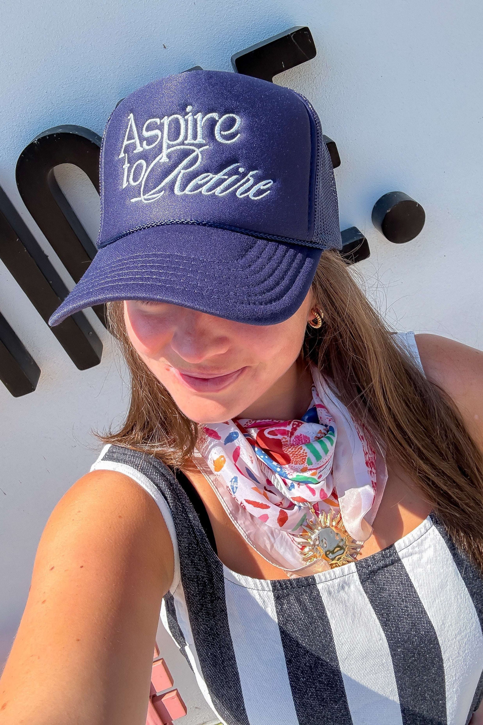 Woman wearing Aspire to Retire Navy Trucker Hat – Foam & Mesh Snapback with white cursive embroidery, styled with striped outfit and floral scarf.