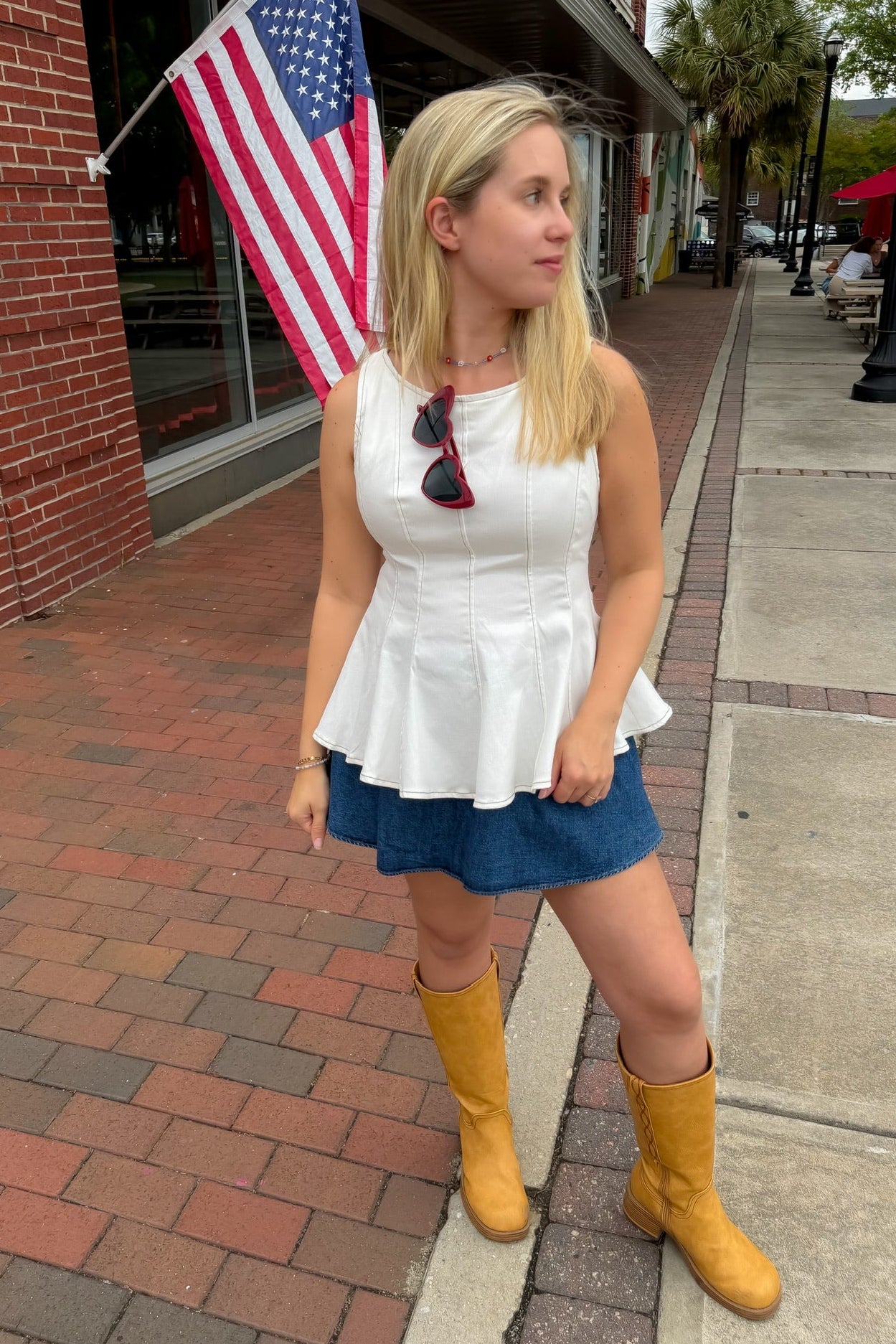 Woman in white top and blue shorts with yellow boots standing on a sidewalk with an American flag in the background.