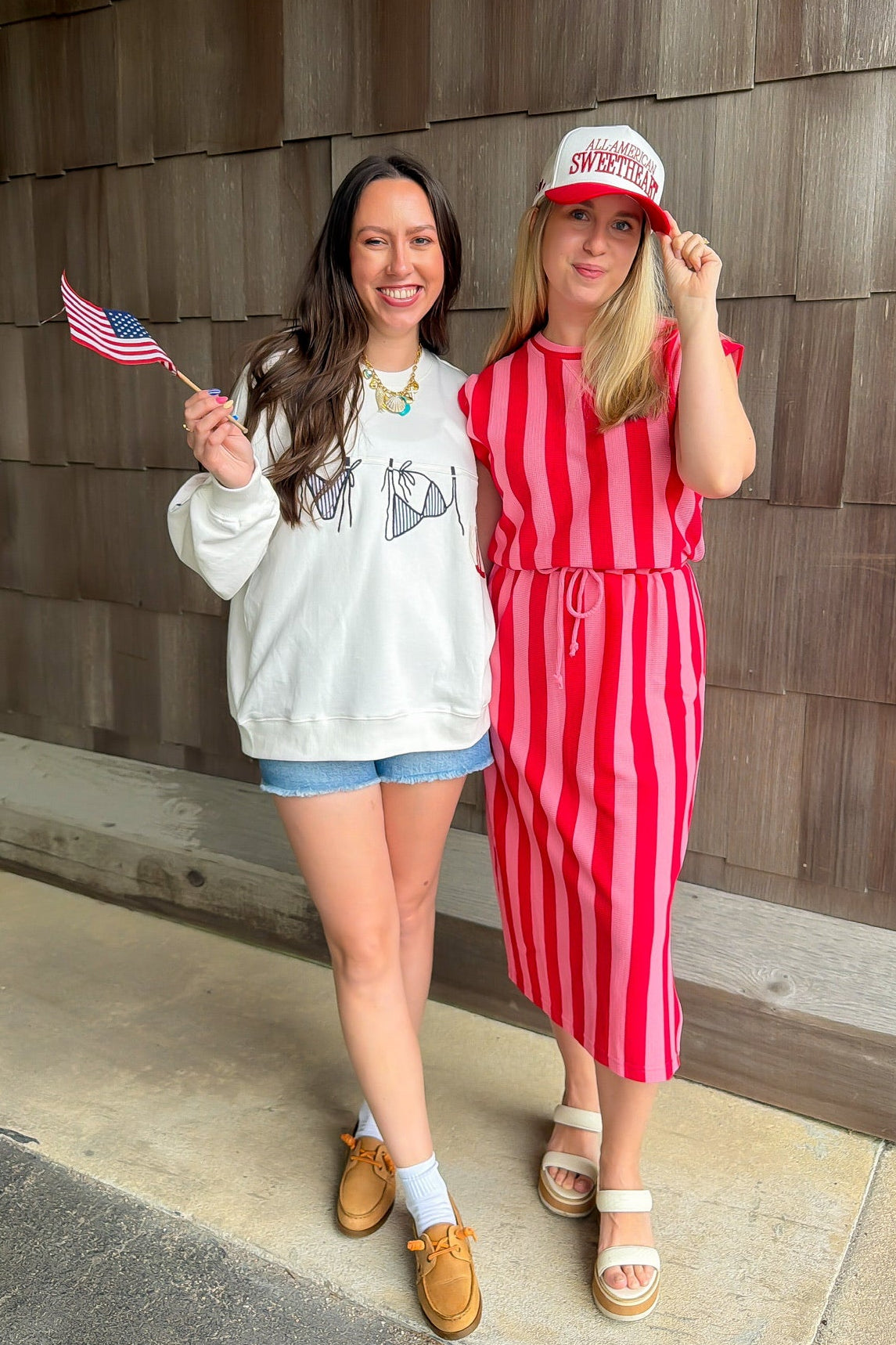 Two women standing outdoors, one holding a small American flag, against a wooden wall.