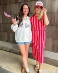 Two women standing outdoors, one holding a small American flag, against a wooden wall.