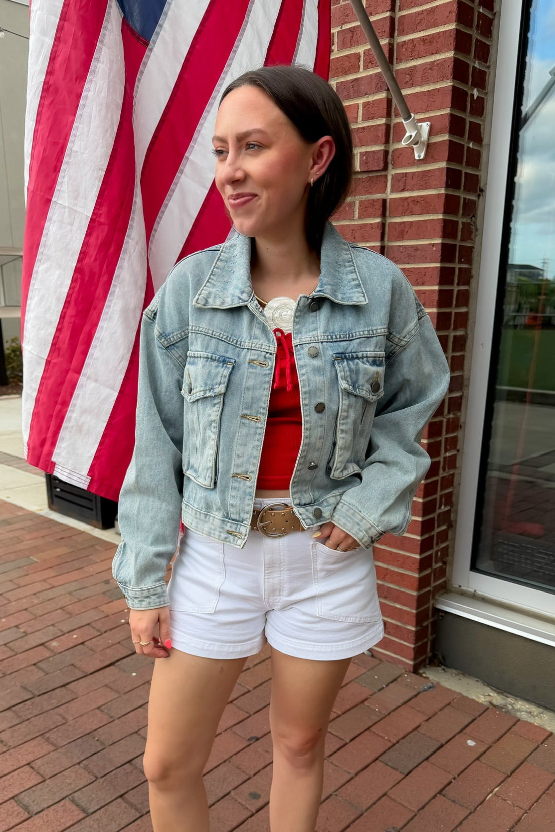 Woman in denim jacket and white shorts standing in front of an American flag.