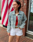 Woman in denim jacket and white shorts standing in front of an American flag.