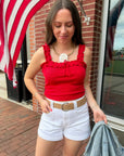 Woman in a red top and white shorts standing outdoors with an American flag in the background
