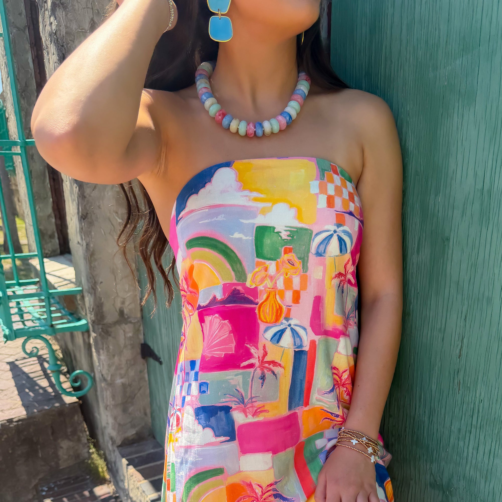 Woman wearing a colorful strapless dress with a pattern of fruits and geometric shapes, standing against a green wall.