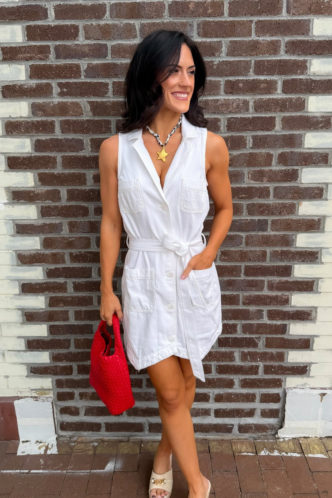 Woman in a white dress holding a red bag against a brick wall.