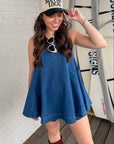 Woman in a blue dress and cap standing next to surfboards against a white brick wall.