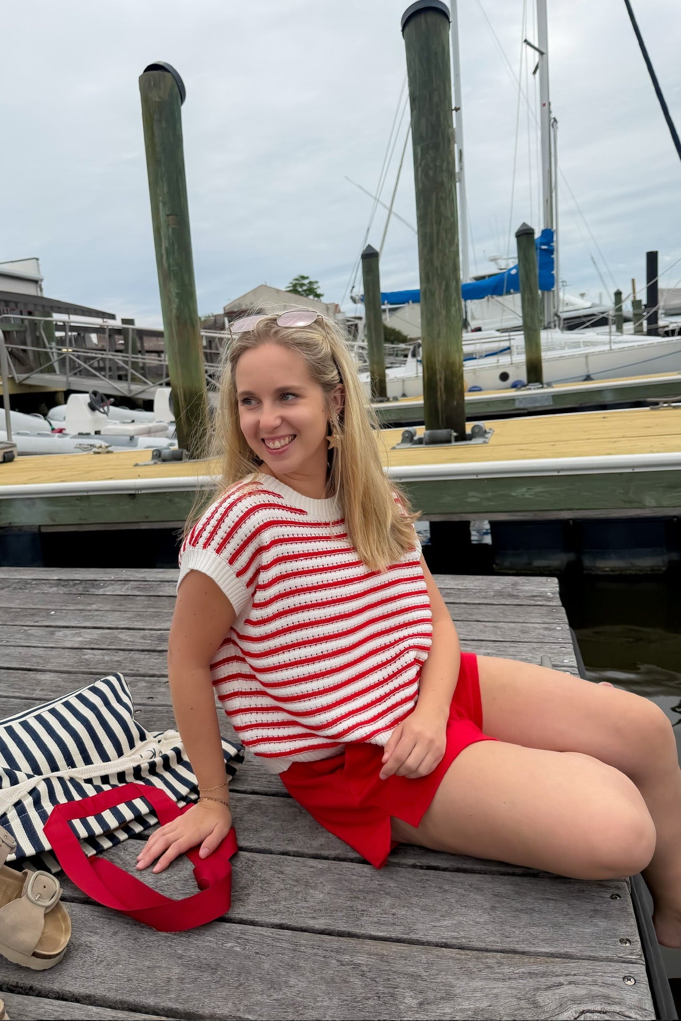 Woman in a red and white striped shirt and red shorts sitting on a dock with boats in the background.