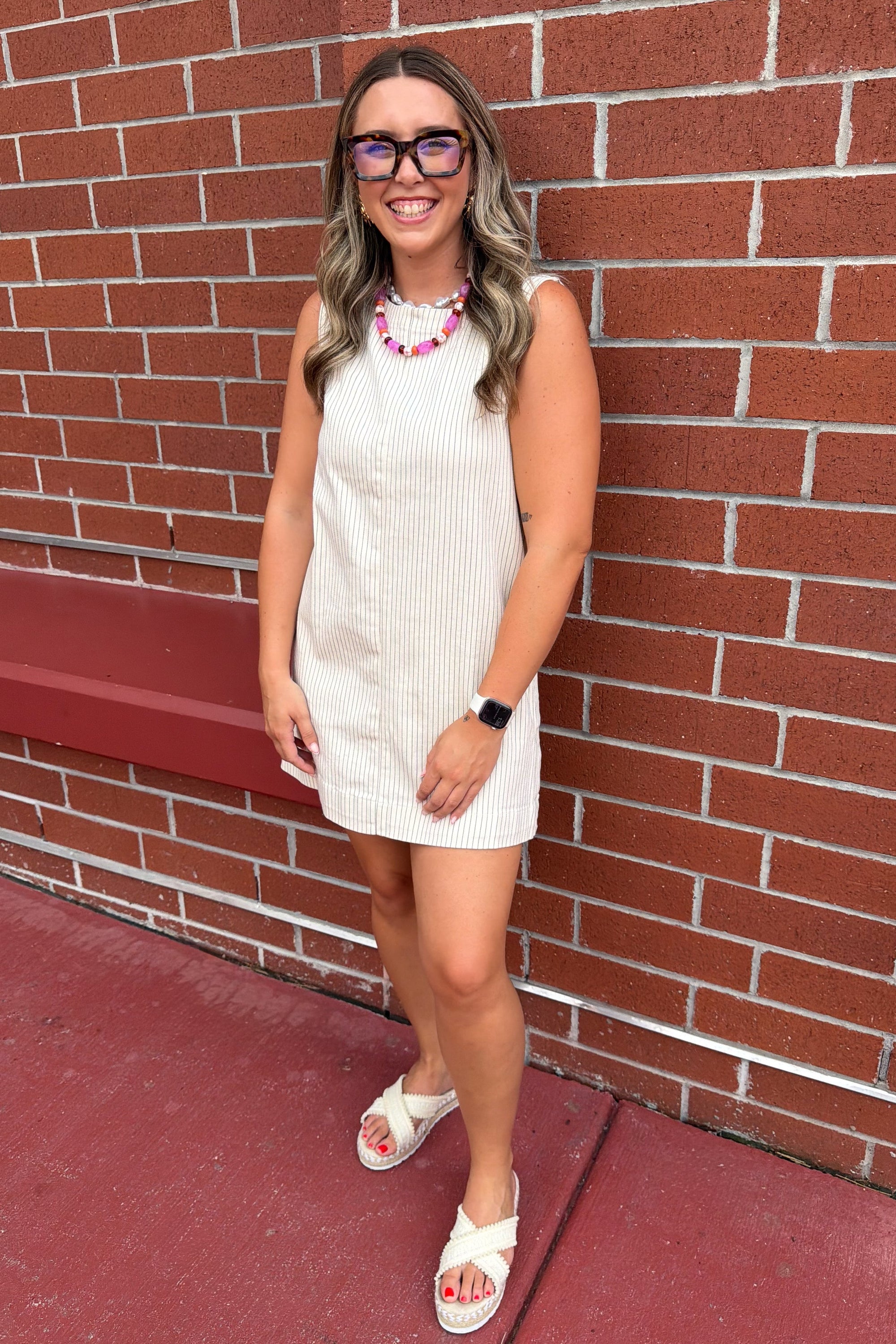 A teacher wearing a sleeveless cream ribbed mini dress, pink and white beaded necklace, and glasses stands against a red brick wall.