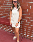 A teacher wearing a sleeveless cream ribbed mini dress, pink and white beaded necklace, and glasses stands against a red brick wall.
