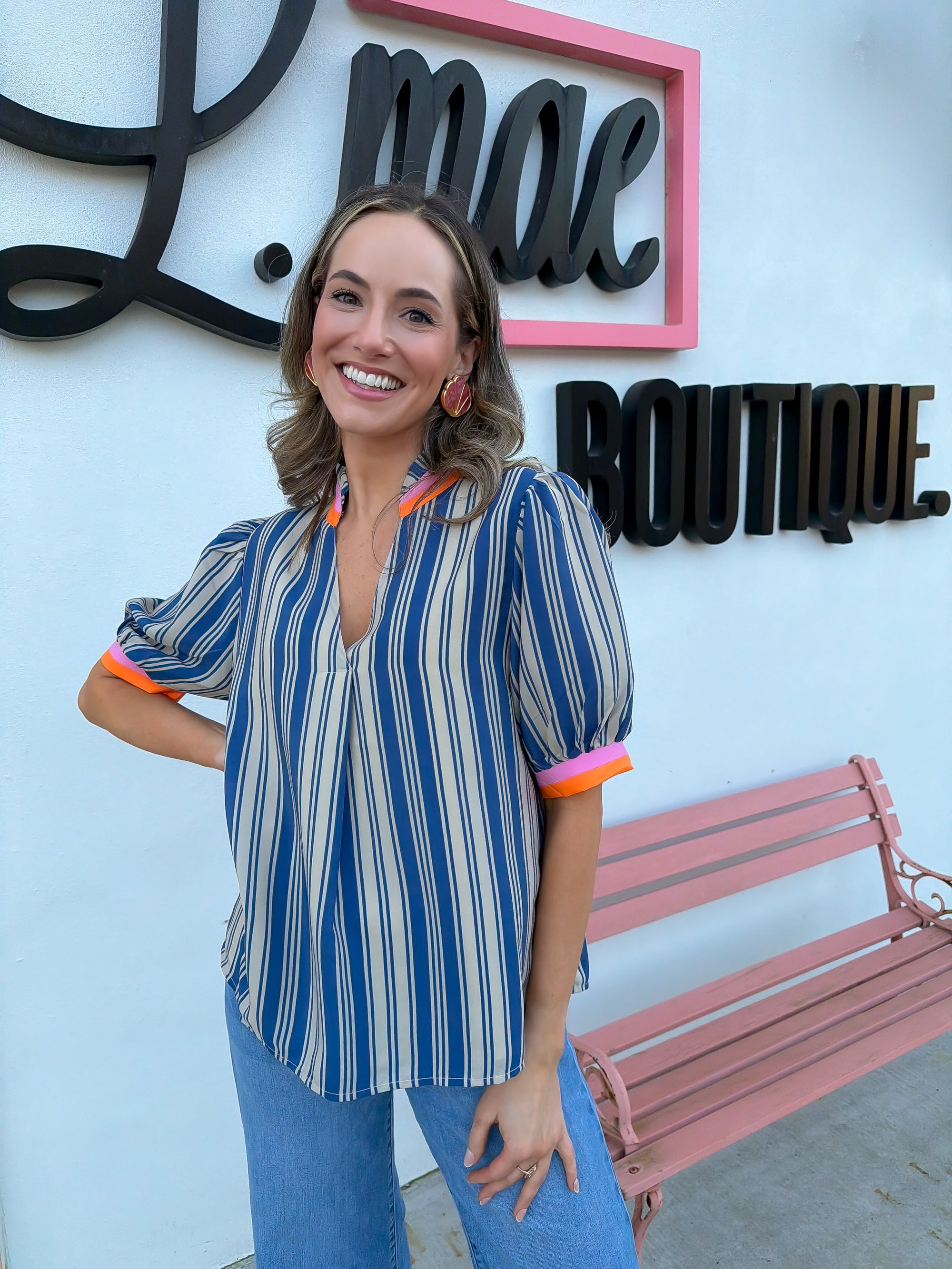 Woman posing in front of a 'L.mae Boutique' sign with a pink bench wearing a THML blouse