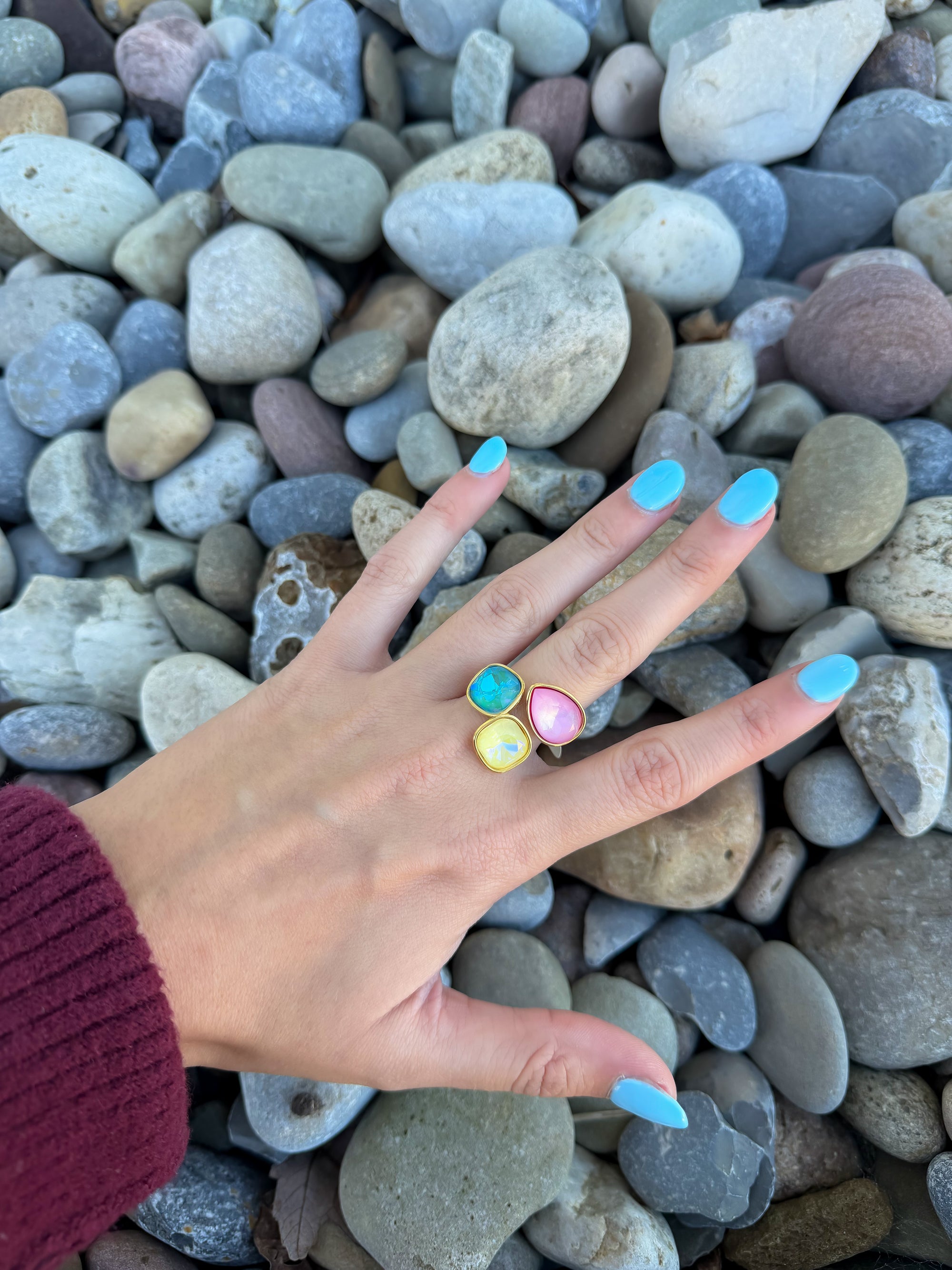 Aurora Gemstone Ring with adjustable open band shown on hand, featuring stackable blue, yellow, and pink stones against a pebble background.