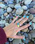 Aurora Gemstone Ring with adjustable open band shown on hand, featuring stackable blue, yellow, and pink stones against a pebble background.