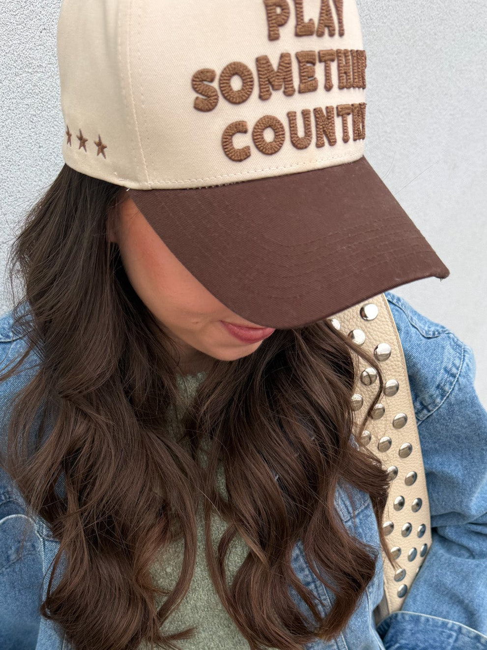 Person wearing a beige and brown cap with 'Play Something Country' text, blue denim jacket, and studded leather bag.