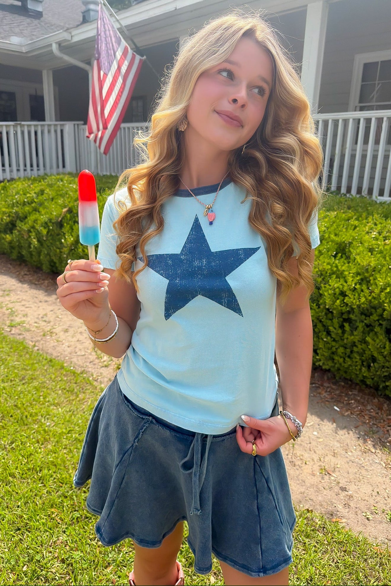 Girl holding a red, white, and blue popsicle  in a baby blue star t shirt in front of a house with an American flag.