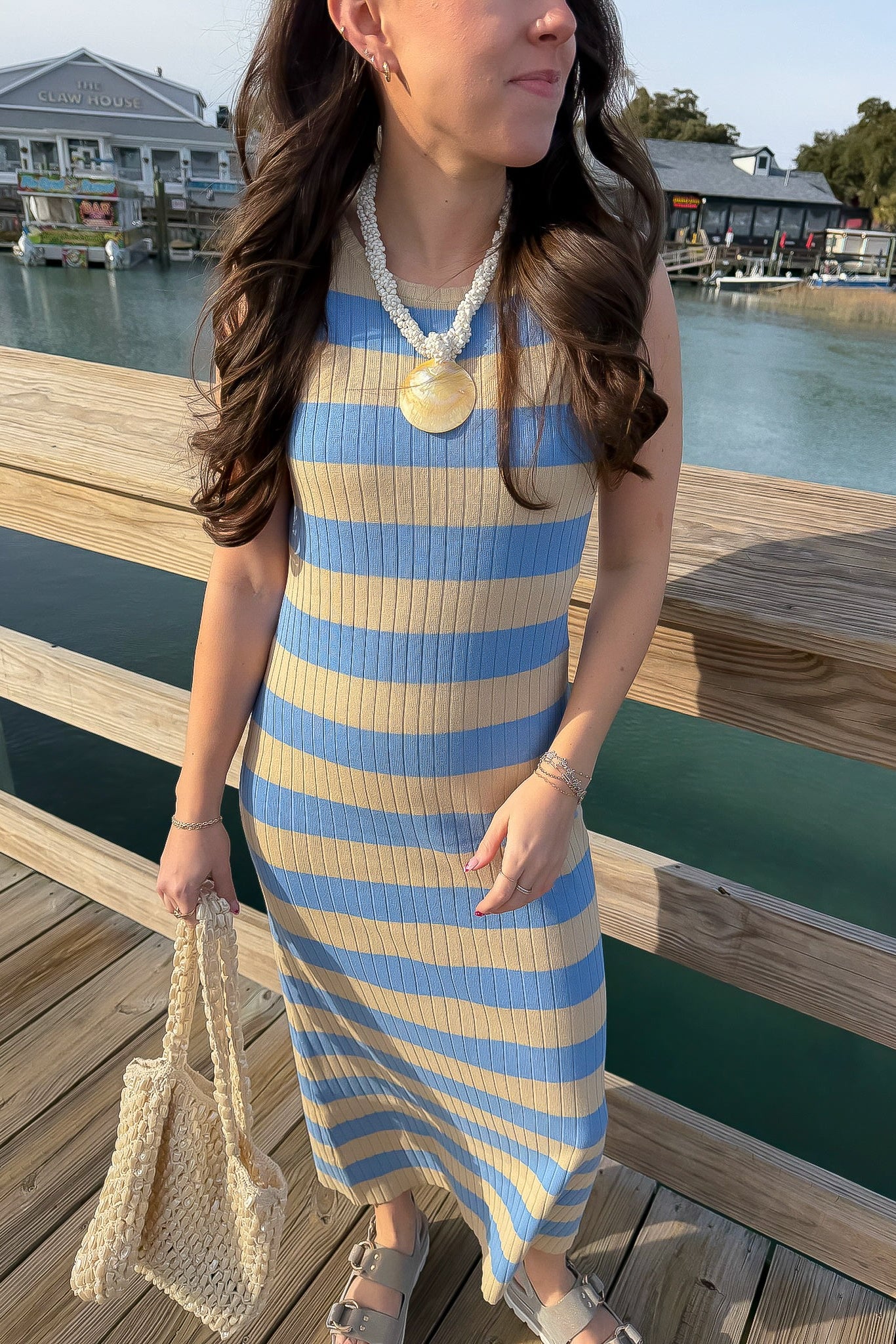 Woman in a blue and beige striped dress standing on a wooden dock by a waterfront.