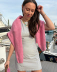 Woman in a white dress with a pink cardigan standing on a dock with boats in the background