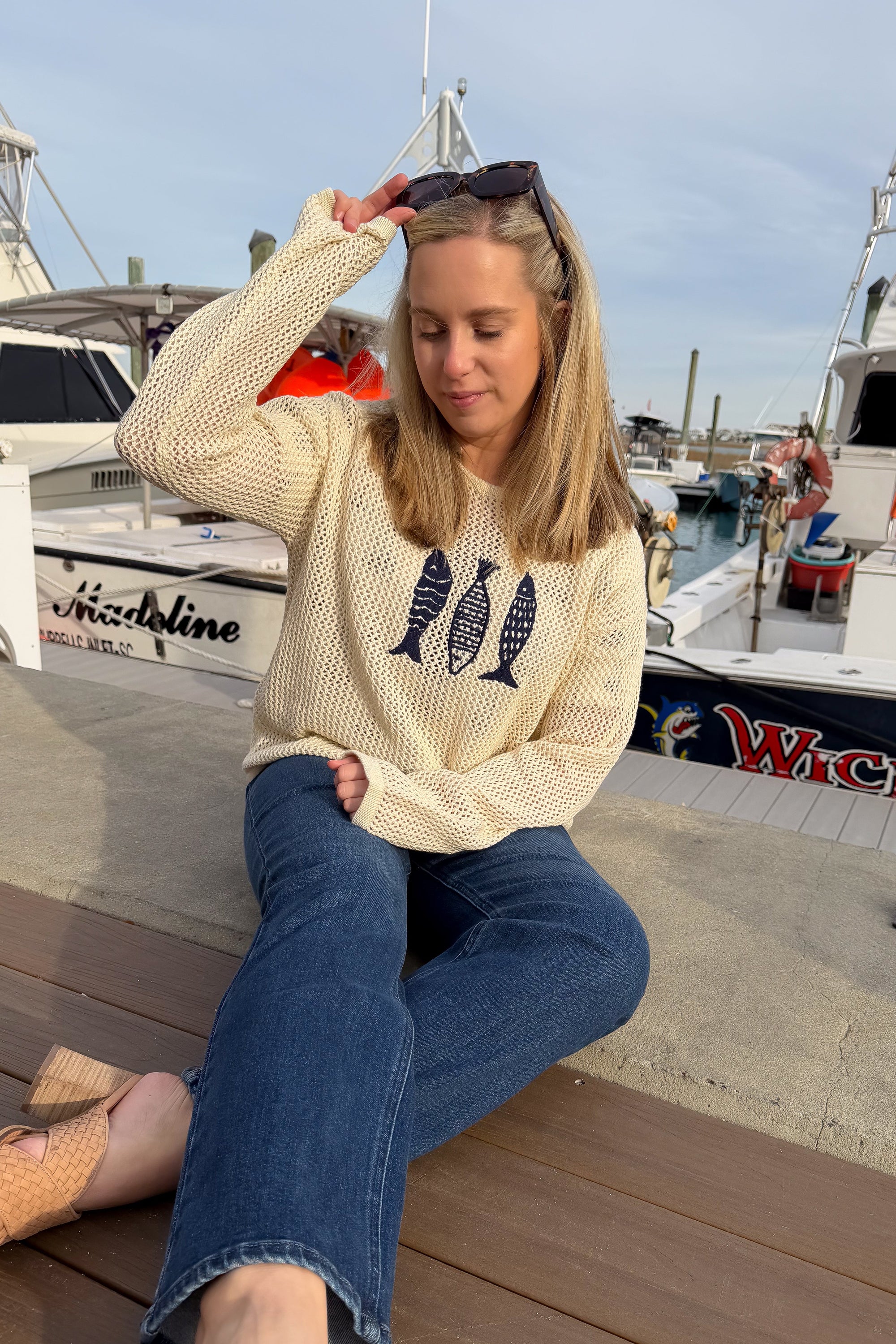 Woman sitting on a dock by boats wearing a beige sweater with fish designs and blue jeans.