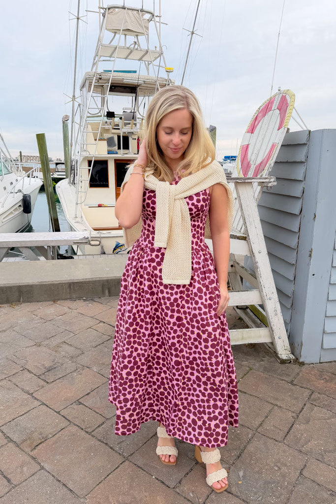 Woman in a pink floral dress with a beige scarf standing on a dock with boats in the background.