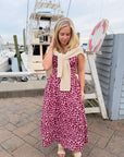 Woman in a pink floral dress with a beige scarf standing on a dock with boats in the background.