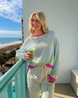 Cabana Stripe Lightweight Sweater in mint and sky blue with pink trim, styled with drawstring pants on a beachside balcony. Ideal spring pullover for casual wear.