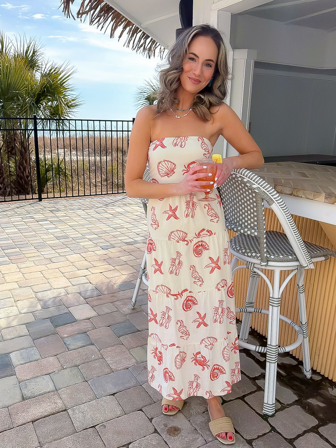 Coastal-chic Shelby Red Seashell Printed Strapless Dress worn at a tropical bar, featuring red shell prints and a relaxed midi fit.
